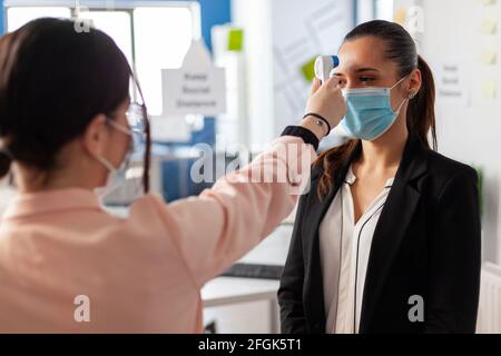 Esame della temperatura corporea del lavoratore in ufficio con termometro digitale durante la pandemia globale con coronavirus. Donna che indossa la maschera facciale come prevenzione di infezione con covid19. Nuova società normale. Foto Stock