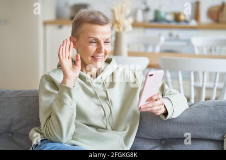 Donna giovane e allegra che svetta con la fotocamera, videochiama con amici o familiari sullo smartphone, siediti sul divano e goditi il tempo libero a casa. Focalizzazione selettiva su donne Foto Stock