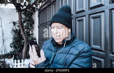 L'uomo anziano effettua una videochiamata con il suo smartphone e. le sue cuffie bianche in giardino Foto Stock