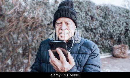 Sorridendo uomo anziano in berretto e cappotto facendo un video chiama con il suo telefono cellulare nero e caschi bianchi dentro il giardino verde Foto Stock