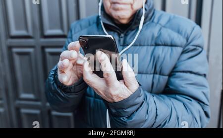 Ritratto delle mani di un uomo anziano che tiene il suo nero intelligente telefono che effettua una videochiamata con le sue cuffie bianche all'aperto Foto Stock