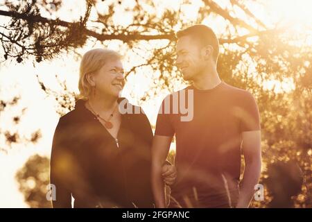 Madre che cammina con il figlio alla luce dorata del sole. Donna anziana e giovane adulto che parlano nel parco pubblico. Foto Stock