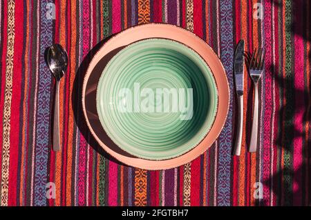 Immagine della vista dall'alto di una piastra e di una piastra per zuppe con posate su una colorata tovaglia a righe al sole Foto Stock