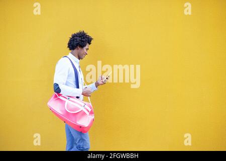 Uomo nero con acconciatura afro che trasporta una borsa sportiva e uno smartphone su uno sfondo urbano giallo. Ragazzo con capelli ricci che indossa camicia e sospenditori Foto Stock