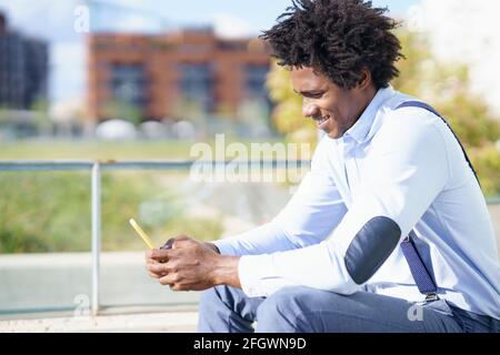 Uomo nero con acconciatura afro che utilizza uno smartphone vicino a un edificio di uffici. Ragazzo con capelli ricci che indossa camicia e sospenditori. Foto Stock