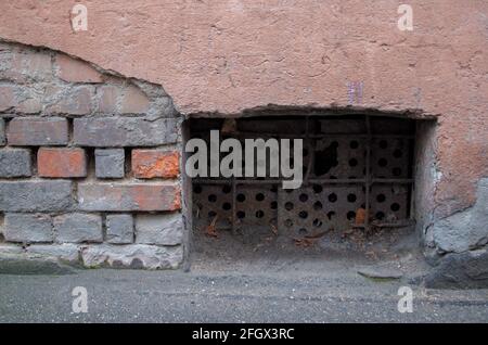 Finestra cava che porta al seminterrato sigillato con lastre in metallo arrugginito forato nella parte inferiore della facciata di una vecchia casa con facciata in rovina, graffiata, fal Foto Stock