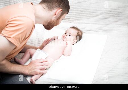 il giovane padre moderno si sta occupando del suo figlio neonato. Cambia il pannolino sul letto. Concetto di igiene e cura del neonato. Amore e cura del Padre. D. Del Padre Foto Stock