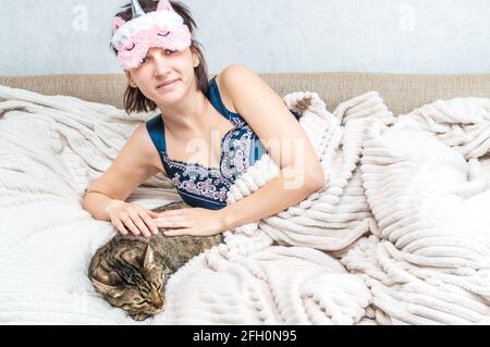 Ritratto di una giovane donna al mattino con una maschera dorme sul viso e pigiami. Nelle vicinanze si trova un gatto grigio che dorme. Concept mattina. Donna è felice Foto Stock