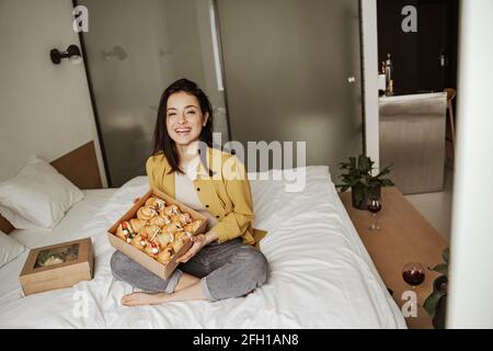 Bella donna seduta sul letto con scatole di cibo pieno di croissant, ha una cena romantica a casa Foto Stock