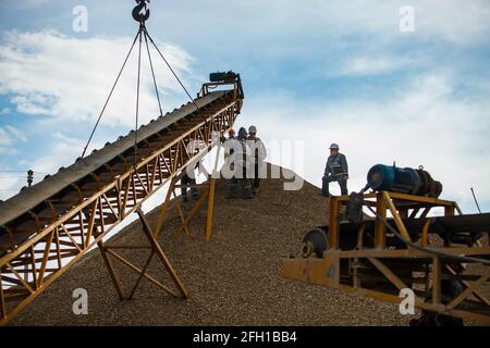 Lavoratori che montano la macchina per frantumare rocce, utilizzando per la frantumazione di minerali d'oro. Ascensori con gru hook.Mining e lavorazione impianti Altynalmas.Blue cielo, nuvole Foto Stock