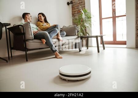 Bella giovane coppia che mostra seduto sul divano nel soggiorno mentre l'aspirapolvere robot funziona. Buona famiglia che si trasferiscono in un nuovo appartamento Foto Stock
