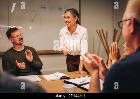 Donna d'affari matura che riceve applausi dalla squadra durante la riunione. gli uomini d'affari si battono per mano di un collega dopo la sua presentazione. Foto Stock