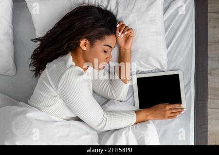 Donna nera sdraiata a letto, dormendo e tenendo la tavoletta Foto Stock