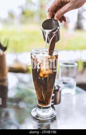 Coltivare barista irriconoscibile versando il latte fresco dal caraffa in vetro di caffè nero freddo servito su un tavolo di vetro all'aperto caffetteria Foto Stock