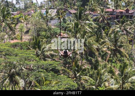 Senza volto brunette ondeggianti su altalene sopra lussureggiante foresta esotica resort Il giorno di sole in Indonesia Foto Stock