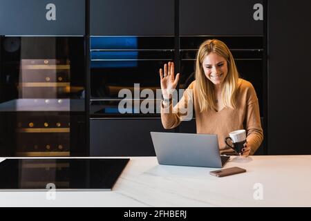 Una donna che sventolava la mano e sorride mentre parla in video chat via computer portatile in cucina a casa Foto Stock
