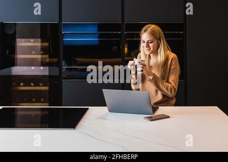 Positivo femmina sorridente che tiene una tazza di tè mentre parla sopra chat video via computer portatile in cucina a casa Foto Stock