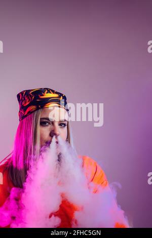Donna cool in stile di strada vestito fumo e sigaretta e. esalare il fumo attraverso il naso su sfondo viola in studio con illuminazione al neon rosa Foto Stock