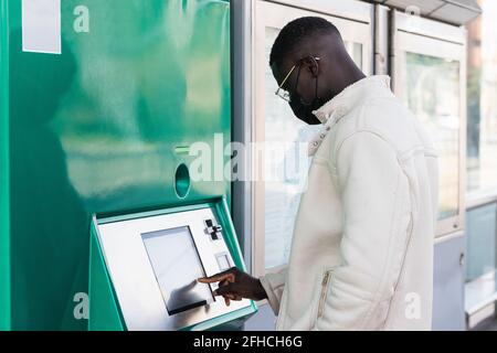 Vista laterale dell'imprenditore maschile etnico in maschera di tessuto utilizzo di una macchina automatica per il teller in città Foto Stock
