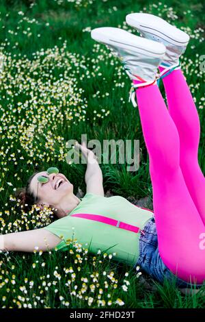 Vista laterale gioiosa giovane donna con outfit luminoso sdraiato erba lussureggiante con gambe sollevate in campagna Foto Stock