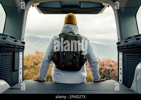 Vista posteriore di un camper maschio irriconoscibile in outerwear seduto sopra furgone e vista panoramica delle highlands Foto Stock