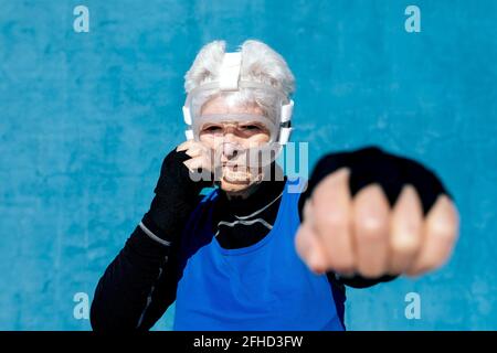Pugile maturo focalizzato in casco e avvolge a mano punzonatura aria contro la parete blu nel centro sportivo all'aperto Foto Stock