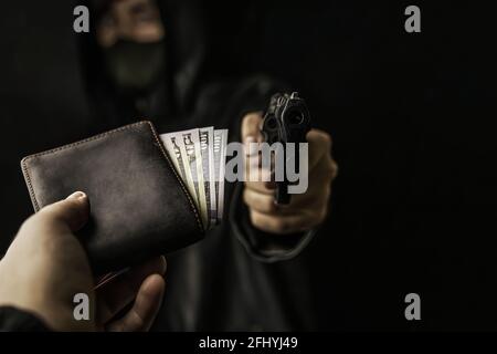 Rapina armata. La mano dell'uomo tiene fuori il portamonete di soldi per derubare con la pistola. L'arma da fuoco è rivolta verso la telecamera. Criminale in cappuccio e maschera. Assalto a un uomo disarmato. Centocinquanta dollari in portafoglio aperto. Foto Stock