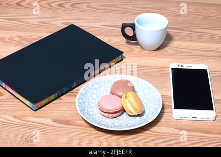 Tazza di caffè bianca servita su un piatto bianco con biscotti macaroon, mobile e colorato taccuino nero su un tavolo di legno. Foto Stock