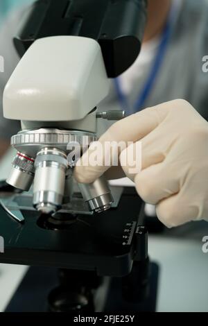 Primo piano di uno scienziato irriconoscibile in guanti che regolano l'obiettivo prima ricerca al microscopio in laboratorio Foto Stock