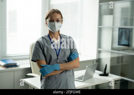 Ritratto di giovane chirurgo sicuro in maschera e guanti in piedi con braccia incrociate contro la scrivania nel proprio ufficio Foto Stock