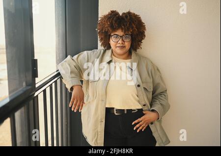 Bella felice sorridente curvy Plus taglia African nero donna afro capelli in posa in T-shirt beige, jeans, occhiali eleganti sul balcone soleggiato. Imperfezione del corpo, concetto di diversità positiva del corpo di accettazione. Foto Stock