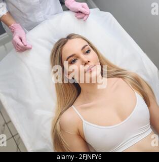 Closeup di mani estetiste in guanti che toccano il viso della giovane donna. Concetto di chirurgia plastica. Bellezza del viso. Ritratto di bella donna bionda con trucco perfetto e pelle morbida e liscia. Foto Stock