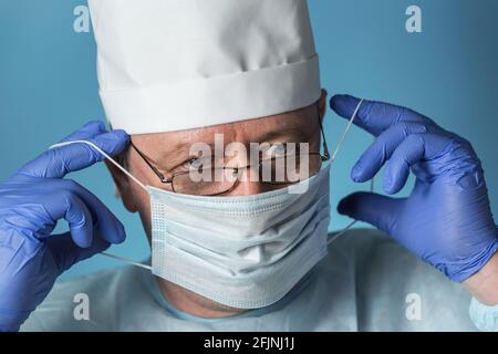 Un medico in abbigliamento medico: Un cappuccio, guanti, vestaglia e occhiali indossa o rimuove una maschera protettiva. Primo piano Foto Stock