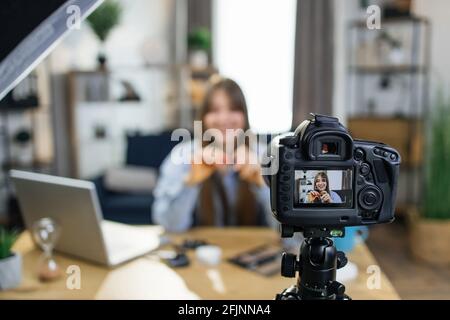 Affascinante giovane donna seduta a tavola con vari cosmetici alla moda e tutorial di registrazione sul trucco professionale. Messa a fuoco sullo schermo della fotocamera digitale. Concetto di blogging. Foto Stock