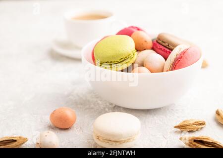 Macaroni multicolore e uova di cioccolato in ciotola di ceramica, tazza di caffè su fondo grigio di cemento. Vista laterale, primo piano, fuoco selettivo, vita morta Foto Stock