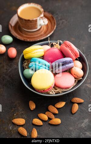 Macaroni multicolore e uova di cioccolato in ciotola di ceramica, tazza di caffè su fondo nero di cemento. Vista laterale, primo piano, vita still. Colazione, colazione Foto Stock