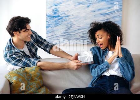 Felice sorpresa giovane attraente african american donna riceve una bella scatola regalo dal suo fidanzato latino per un compleanno o relazioni anniversario, guarda il regalo, le aggrappa le mani Foto Stock