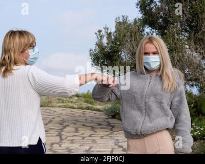 Due donne con maschere davanti ai loro volti, in piedi lontani, si salutano con pugno a pugno, il 'Corona greeting', all'aperto Foto Stock