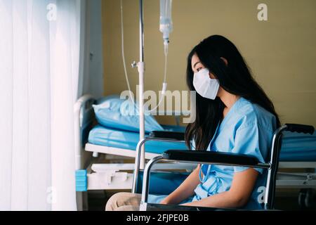 Triste e sconvolto giovane paziente con maschera chirurgica seduta premuto sulla sedia a rotelle nella stanza dell'ospedale accanto alla finestra Foto Stock