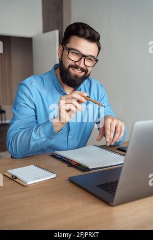 Giovane uomo felice positivo sorriso parlare al computer portatile fotocamera sedersi alla scrivania dell'ufficio domestico Foto Stock