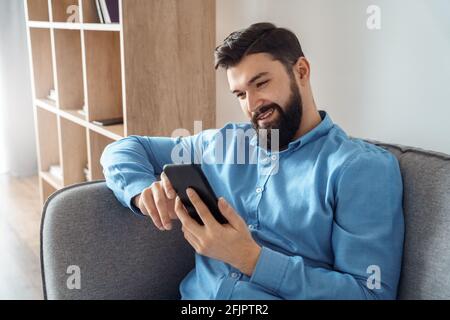 Uomo caucasico sorridente che utilizza un dispositivo smartphone per rilassarsi a casa divano Foto Stock