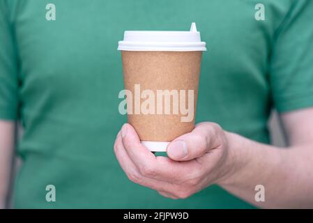 Un uomo in t-shirt verde che tiene la tazza marrone di carta con bevanda - caffè o tè, tazza per il take-away, mockup con spazio per il testo Foto Stock