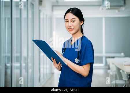 Ritratto di giovane bella dottoressa asiatica in uniforme blu con stetoscopio in piedi nel corridoio dell'ospedale che tiene gli appunti di controllo dei referti mentre si guarda la fotocamera Foto Stock