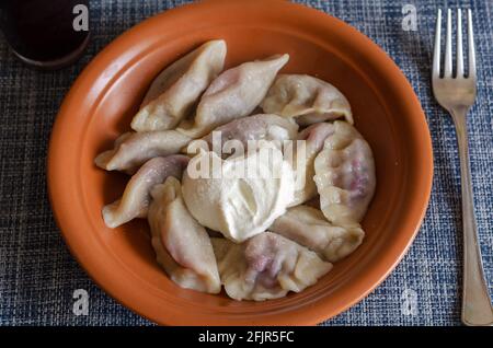 Concetto di cucina tradizionale Ucraina. Varenyky con ciliegie. Una porzione di gnocchi pronti da mangiare in un piatto di argilla marrone con panna acida, cosparsa wi Foto Stock