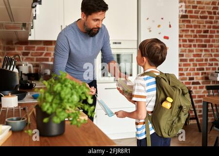 Padre che dà il suo figlio pranzo scatola e maschera protettiva viso Foto Stock