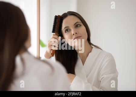 Ragazza in accappatoio spazzolando i capelli marroni lisci allo specchio Foto Stock