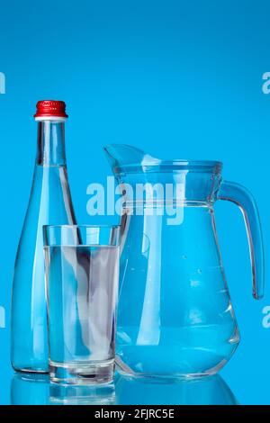 vetreria riempita con acqua potabile pulita per oggetti sanitari vetro e caraffa con manico e bottiglia con coperchio ancora vita su sfondo blu. Foto Stock