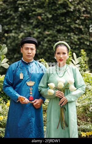 Ritratto di una giovane coppia vietnamita preoccupata in costumi tradizionali situato nel parco cittadino con fiori di loto e fan della carta Foto Stock