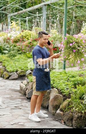 Sorridente giovane serenhosue vietnamita che parla al telefono e controlla fiore viola petunia in vaso sospeso Foto Stock