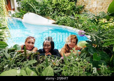 Gruppo multietnico di giovani donne felici che trascorrono del tempo a nuotare piscina dell'hotel termale decorata con splendide piante lussureggianti Foto Stock